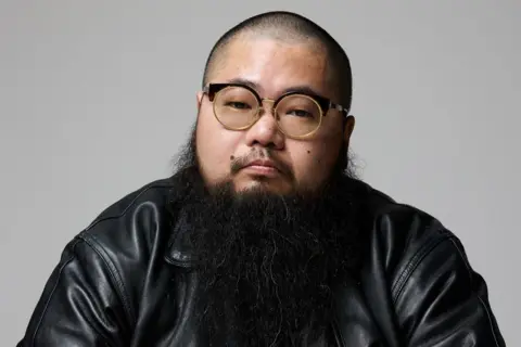 BBC/Supplied: Badiucao A man with a shaved head and a long black beard, wearing wide-rimmed glasses and a black leather jacket, looks into the camera