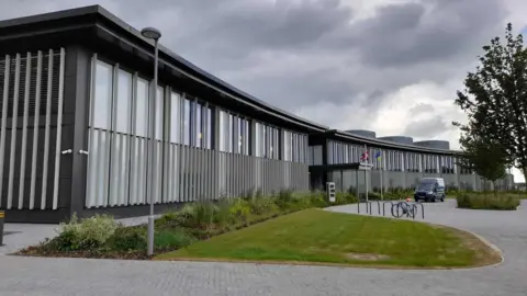 A modern office building with shrubs planted outside, grass and a paved area. Flags, including the union flag and Ukraine flag, can be seen flying from flagpoles outside. A van and bicycle are parked nearby.