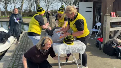 Matt Bailey, wearing a grey t-shirt and yellow and black bobble hat, lies face down on a massage table by a lake being massaged by three women dressed in black t-shirts and yellow vests. A laughing Anna Foster, with blonde hair and wearing black headphones, holds a microphone under the bed to interview him.