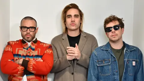 Mark Allan/BBC The pop-punk band Busted looking straight at the camera. Matt Willis is wearing black glasses and a red patterned jacket with his arms folded. Charlie Simpson is wearing a black top and beige coat. James Bourne is wearing a green top, denim jacket, and black sunglasses.