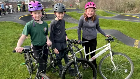 Gavin Kermack / BBC Three children - a girl, a boy and another girl, all around 10-11 years old, stand in front of the pump track. They are wearing cycle helmets and are holding bikes.