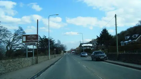 Colin Pyle/Geograph Photo of Ffordd Holway in Holywell in Flintshire. Cars are visible parked on the curb and there's a sign for Holywell/ Treffynnon on the left. There are houses on the right hand side. 