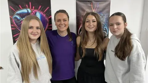 Four women standing together, all looking into the camera and smiling. All have long hair, two wearing hoodies, one a T-shirt, another a dress.