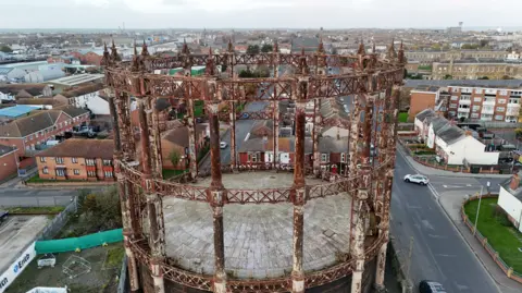 Drone image shows rusting gasometer structure rising over nearby homes and businesses.