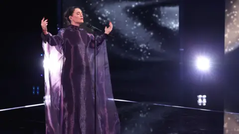 Getty Images Jessie Ware is on-stage at the Bafta Film Awards, wearing a long, purple dress. Its fabric hangs from her open arms as she singers into a microphone. Her eyes are closed as she sings, with white light illuminating her dress.