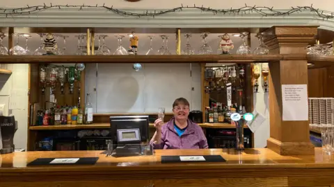 Sally Fairfax / BBC A wooden bar. A woman is standing behind the bar holding up a glass. Various drinks can be seen behind her and there are a couple of beer pumps on the bar. 