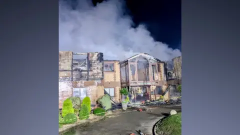 Essex County Fire and Rescue Service The front of the clubhouse building at West Essex Golf Club, heavily damaged by fire. The photo is taken at night with the building illuminated by light, and grey smoke rises from the building, which has an apex above the main entrance and several burnt-out window frames. There is tape running across the front of the building.