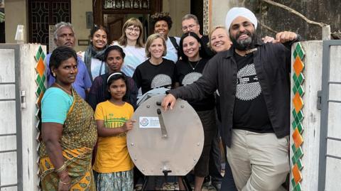 A group of people stand alongside a hand-cranked washing machine