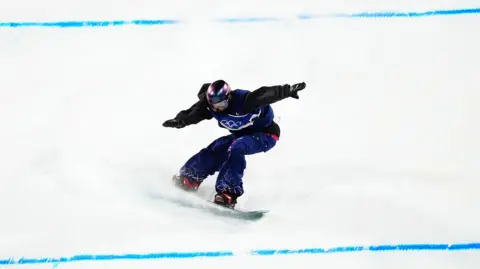 PA Media Mia Brookes wearing a Team GB snowboarding outfit, including a purple helmet, has her arms outstretched as she lands a trick on a snow slope. There are two blue lines across the white slope.