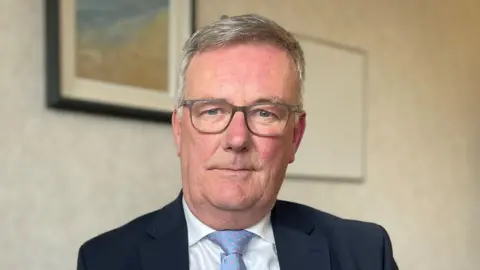 PA Media Mike Nesbitt wears a black suit, white shirt and blue tie. He is wearing glasses and has grey hair. He is looking at the camera. There are photos on the wall behind him. 