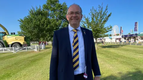 BBC/Seb Cheer National Farming Union (NFU) president Tom Bradshaw. He wears glasses, a dark blue suit and a white shirt with a yellow and blue tie. He stands in a field, with a green tractor in the corner of the shot.