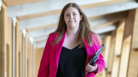 PA Media Gillian Mackay walks to the enclosure  successful  the Scottish Parliament, carrying a achromatic  folder. She is wearing a agleam  magenta jacket.