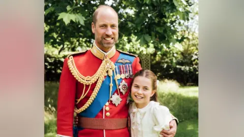 Prince William is wearing the scarlet dress uniform of the Welsh Guards, of which he is colonel, and has his arm around Charlotte, who is wearing a cream dress.