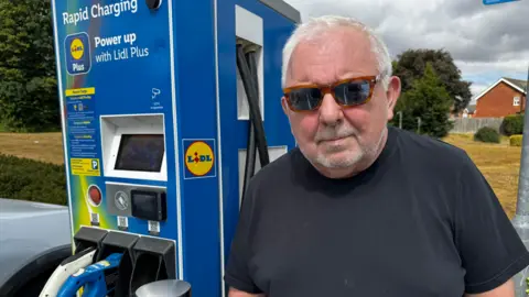 A man wearing sunglasses and a black T-shirt stands beside a blue Lidl Plus rapid electric vehicle charging unit at Lidl's Barton-upon-Humber store. Charging cables are connected to a vehicle parked alongside the charger. The unit displays instructions for using the charging service, while trees, housing and dark clouds are visible in the background.