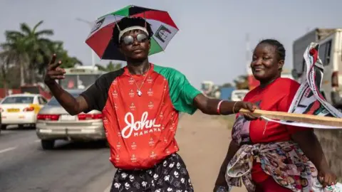 AFP Two supporters of John Mahama pose by the roadside for a photograph in Accra on 3 December 2024. One is wearing the emblem of the National Democratic Congress - an umbrella - as a hat