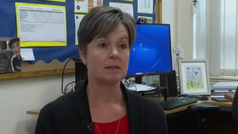 Sue Julyan wearing a black blazer, red top underneath and silver necklace sat in front of a computer on a desk which also has a colourful piece of artwork on it. Behind her on a board is a photo of a smiling young man, possibly a teenage relative of hers. Sue has a short haircut with dark hair tinged with grey.