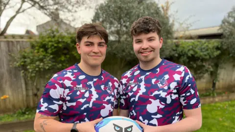 Two people wearing matching pink, blue and white camouflage rugby shirts stand on grass in a garden, holding a rugby ball with a stylised black‑and‑white face logo on it.