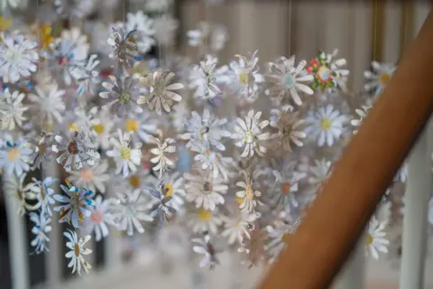 Gillian Taylor A curtain of paper daisies hangs in the stairwell. Some are in focus, others are blurred in the background. There is visible handwriting across the petals of the daisies. 