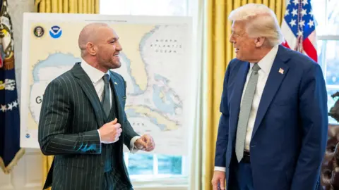 Conor McGregor is wearing a green pin stripe suit and is laughing. He has short cropped hair and stubble.  Donald Trump, who is wearing a navy suit and has blond hair, is looking at McGregor.