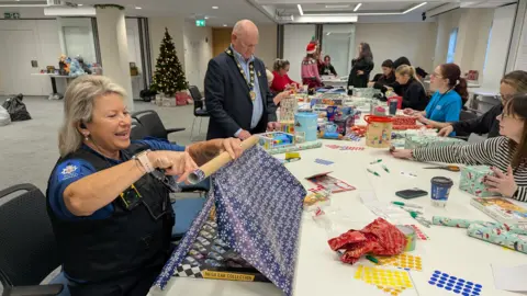 A woman in police uniform wrapping a present on a long, white table with several volunteers in the background doing the same