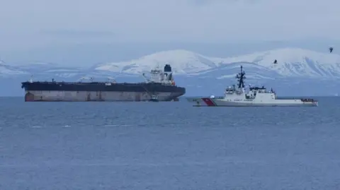 The tanker is a large ship with rusty marks on its hull. The upper part of its hull is painted blue and it has a white bridge and a single blue funnel. In the foreground is a white US Coast Guard vessel. There are snow-covered hills in the background.