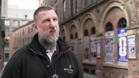 A man in his 40s, with short light brown hair, a grey beard and a large tattoo on his neck, speaks to the camera in front of a large brick building lined with posters. He is wearing a black zip-up fleece.