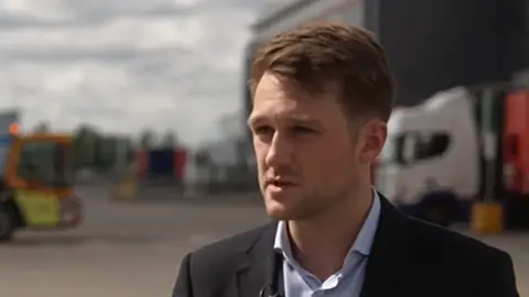 A young man wearing a suit giving an interview at a lorry depot