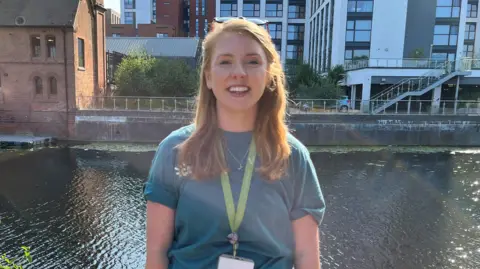 Juliette Burt smiling at the camera wearing a turquoise top and a lanyard. She is stood in front of the river