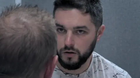 Channel 4 A still of the episode of 24 Hours in Police Custody where Adam White, with short hair and a beard, is looking concerned during a police interview