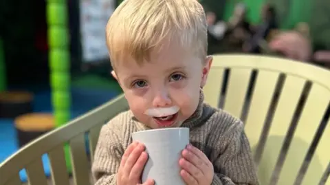 Family handout Hudson is wearing a brown jumper and drinking from a cup and has what looks like milk left on his top lip. He has blond hair and is sitting in a light green chair