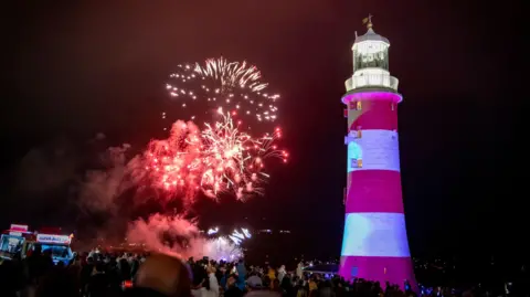 A number of fireworks explode behind Smeaton's Tower. The tower is red and white striped with a lantern at the top.