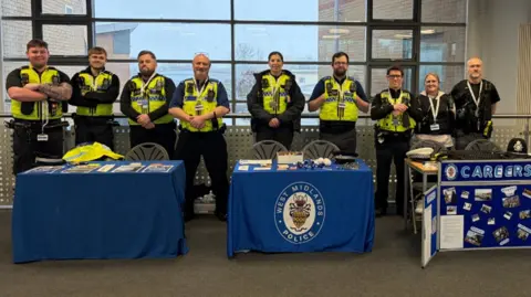 Nine West Midlands police staff members line up behind blue tables and in front of a window. Two of the tables are covered in blue table clothes and a second behind a careers pinboard. Seven are wearing police uniform and yellow high-vis jackets with walkie talkies attached. Two pictured are women.