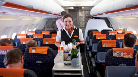 Interior of an Easyjet plane cabin showing female air steward in easyjet uniform pushing a drinks trolley forward and smiling. Also pictured are the back of passengers heads on either side of the aisle 