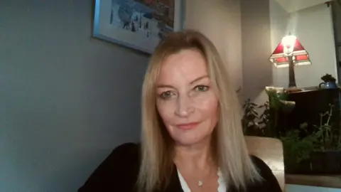 Katherine Hart A woman with long blonde hair looks at the camera, she is wearing a black top and is in room with plants and a lamp behind her