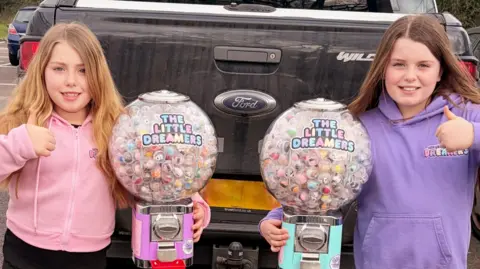 Two girls in pink and purple hoodies with thumbs up, beside two of their spherical vending machines
