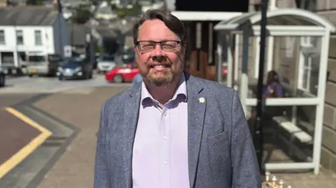 Dan has brown hair and beard, with glasses, and wears a grey jacket and light coloured shirt. He is standing in front of a bus stop in Bodmin town centre. 