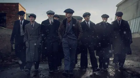 Netflix A film still showing nine men walking while dressed in coats, suits and hats. It looks like snow is falling around them. The colours are dark and muted. Buildings are either side of them. 