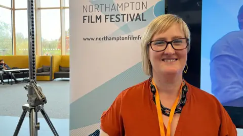 Becky Carrier with medium-length blonde hair wearing dark-framed glasses and an orange short-sleeved top. She also has an orange lanyard and is smiling for the camera. She is standing next to a blue and white Northampton Film Festival banner. There is a lighting stand to the left and tall windows beyond.