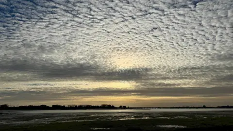 It is nearing sunset and the sky is darkening but is still filled with a multitude of clouds that look like balls of cotton wool. Underneath the sky is an expanse of water with several trees silhouetted against the horizon.