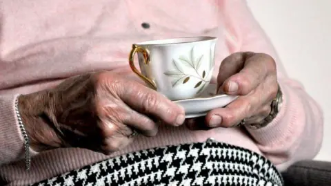 Elderly woman's hands holding a cup and saucer, she is wearing a pink top and a black and white skirt.