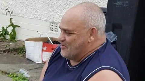 Police Scotland A man with short white hair and a dark blue vest looking off camera while sitting in front of a white wall.