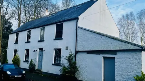 The image shows a white-painted building surrounded by tall leafless trees. The structure is a two‑storey property with a slate roof and a prominent chimney. The main section has several evenly spaced windows and a central door, all framed in dark trim. Attached to the right side is a single‑storey extension with its own door and a slightly sloping roof. A dark-coloured car is parked on the road in front of the building. 