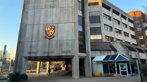 Civic offices at Woking Borough Council. The words 'Woking Borough Council' can be seen on the front of the grey concrete building, as can a red and yellow coat of arms. A person walks next to the steps which lead to the entrance.