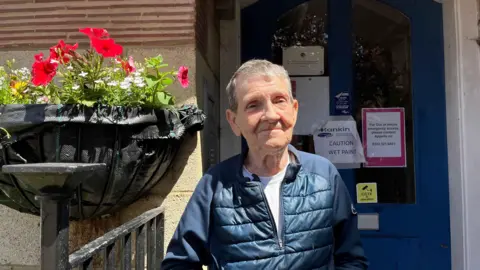 George Carden/BBC Chris Parrish wearing a jacket outside the front door of the flat which is blue. Next to her is a hanging basket of flowers
