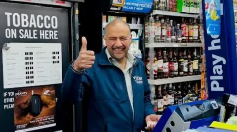 A bald man earing a grey shirt and blue zip-up jacket smiles widely and gives a thumbs up to the camera. He is standing behind a till in a convenience store with shelves of drinks behind him.