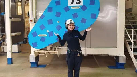 Maria is wearing a white helmet and dark purple hijab and wearing blue long sleeve top that has MARS written on the side and blue trousers and keys and accessories hooked on the belt. She is standing behind a tank in a factory. 