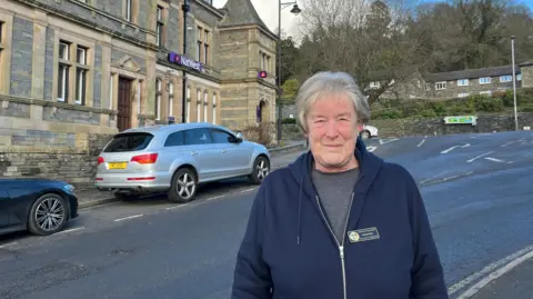 Christine Hallatsch standing outside NatWest in Windermere. She has white hair in a short bob and is wearing a blue hoodie over a grey T-shirt.