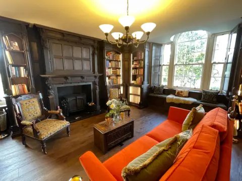 Wood-panelled sitting room with a carved fireplace and built-in bookshelves filled with books, featuring a red sofa, upholstered armchairs, a central wooden coffee table with flowers, a chandelier overhead, and a large bay window with cushioned seating letting in daylight.