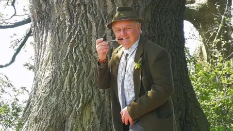 Rob McBride Rob McBride is standing in front of the Darwin Oak tree. He is wearing a hat, shirt, and suit jacket, and is holding a pipe to his mouth