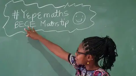 Angela Tabiri Dr Angela Tabiri melihat ke arah dan menunjuk ke papan tulis dengan beberapa tips matematika yang ditulis dengan kapur selama pelajaran YouTube FemAfricMaths untuk siswa sekolah menengah pertama